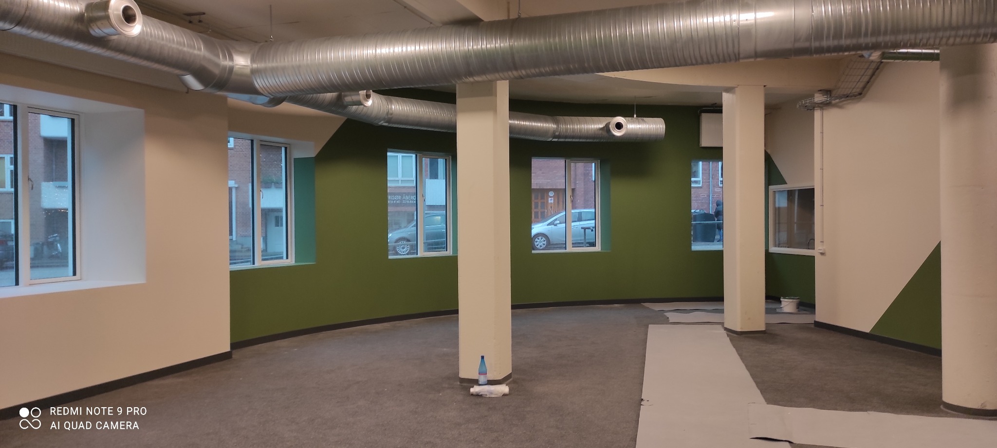 Wnętrze komercyjne po świeżym pomalowaniu ścian na biało i oliwkowo-zielono, z widocznymi kanałami wentylacyjnymi pod sufitem i przygotowanym podłożem pod nową podłogę.