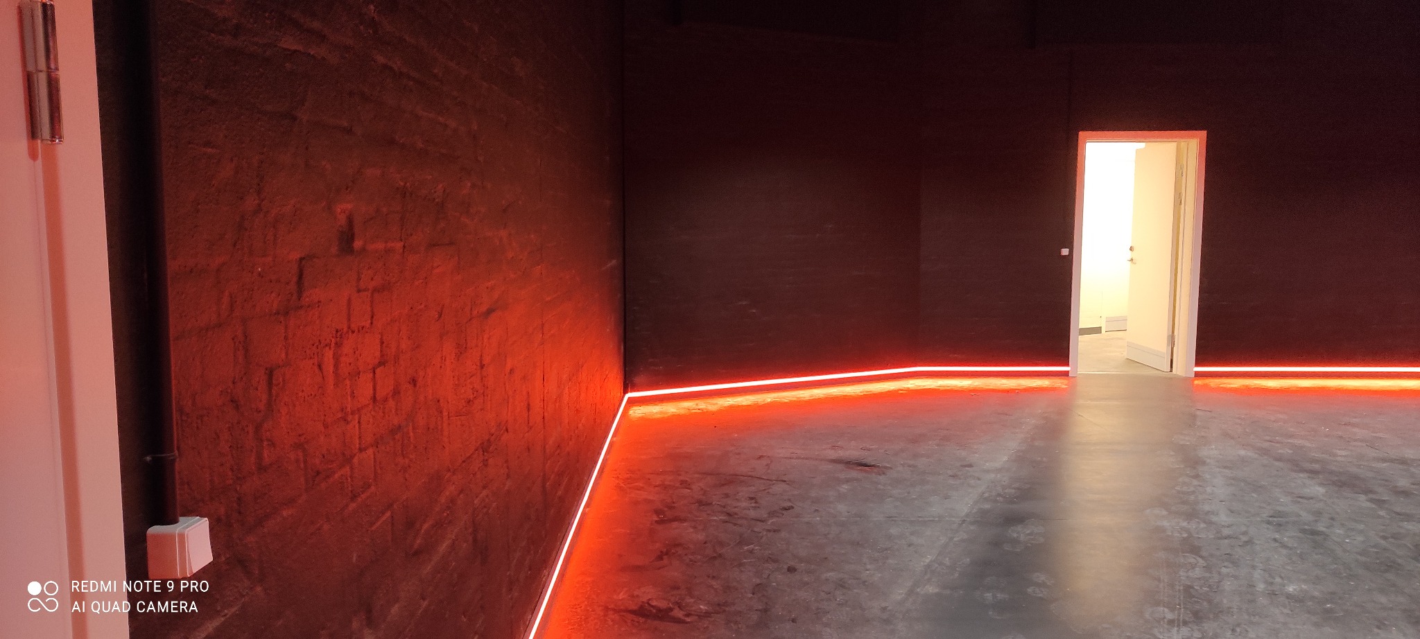 Surowa ściana z cegły pomalowana na ciemny kolor, oświetlona poziomą linią czerwonego światła LED, widoczny fragment drzwi po prawej stronie.