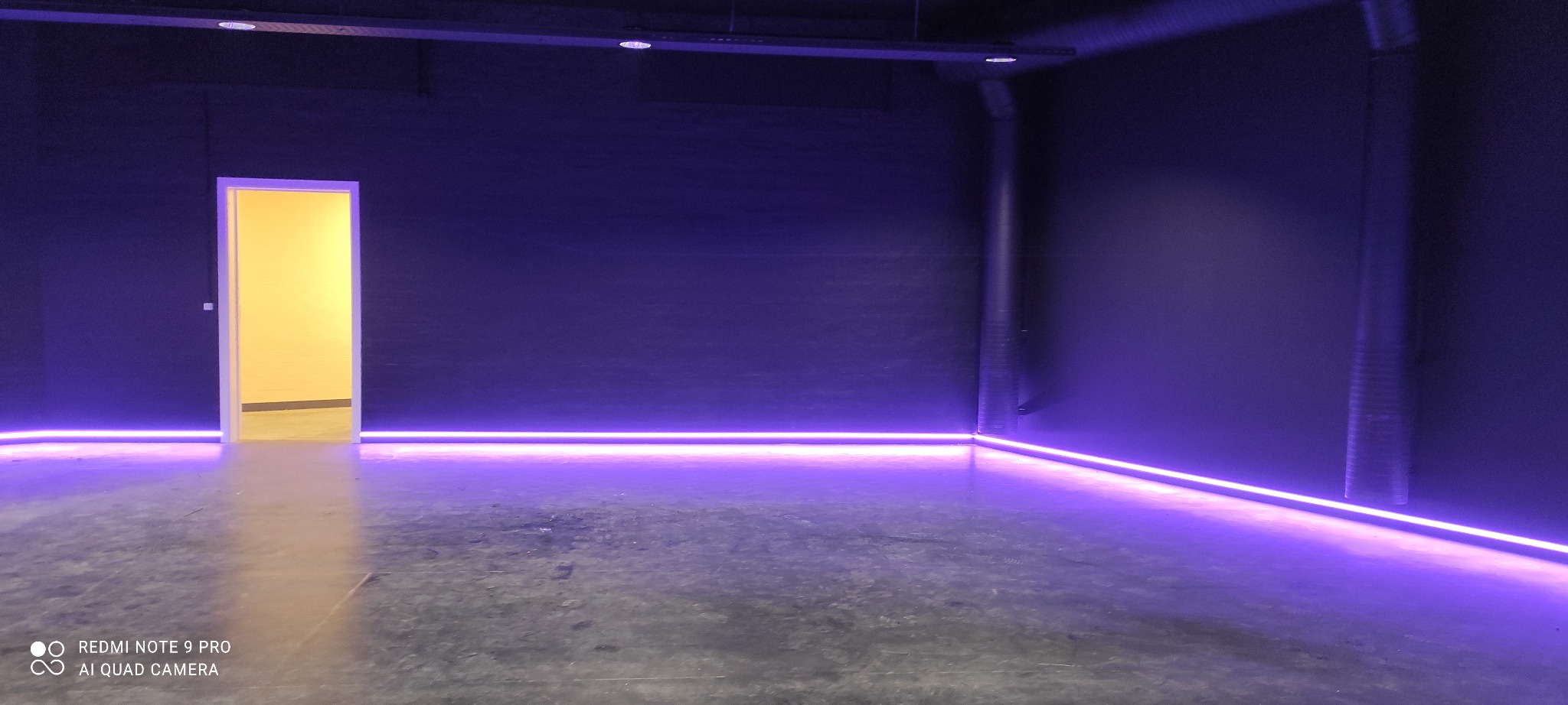 Pomieszczenie z ciemnymi ścianami i podświetleniem LED wzdłuż podłogi, widoczny fragment rury przy ścianie i otwarte drzwi z żółtym światłem w tle.