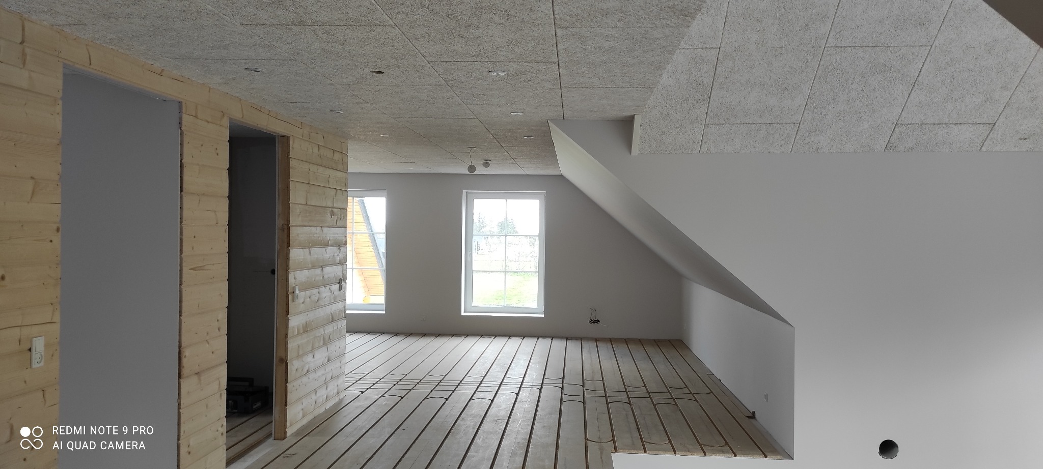 Wnętrze poddasza w trakcie prac wykończeniowych: widoczne drewniane panele podłogowe z ogrzewaniem podłogowym, ściany częściowo obłożone drewnem, sufit z płyt z widocznymi punktami oświetleniowymi.