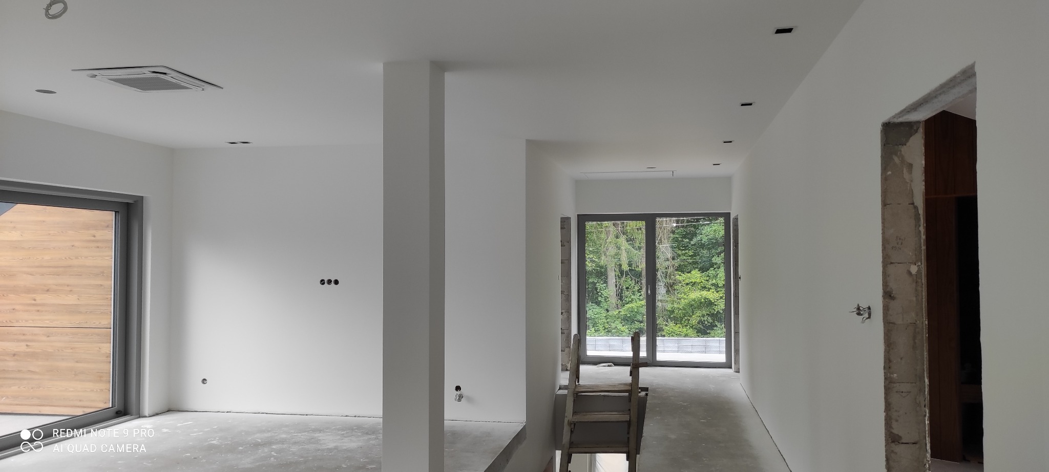 Wnętrze w trakcie remontu: białe, świeżo pomalowane ściany, surowy beton na podłodze, widoczne instalacje elektryczne, schody drewniane prowadzące w dół, okno z widokiem na zieleń.