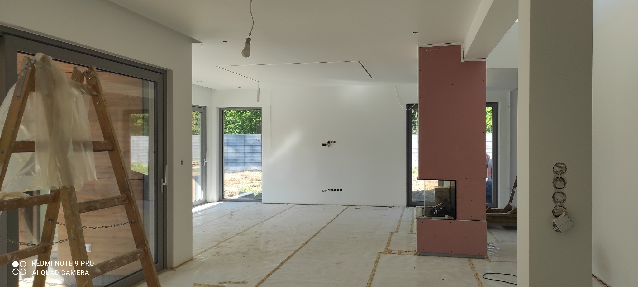 Wnętrze w trakcie prac wykończeniowych: zabezpieczona folią podłoga, ściany przygotowane pod malowanie, widoczna drabina malarska i obudowa kominka z płyt gipsowo-kartonowych.