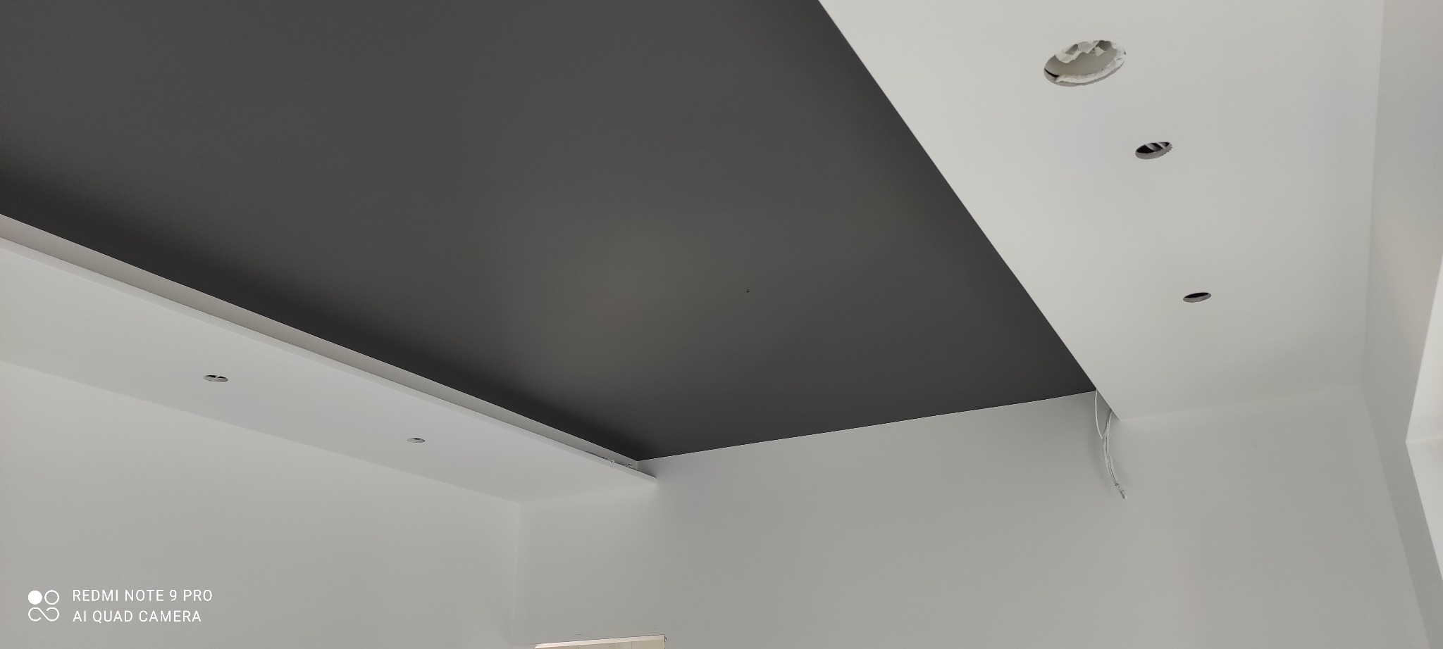 Częściowo pomalowany sufit w kolorze grafitowym z widocznymi otworami na oświetlenie punktowe i wystającymi przewodami.