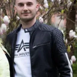 Młody mężczyzna w czarnej skórzanej kurtce z pikowanymi ramionami, stojący na tle kwitnącej magnolii.