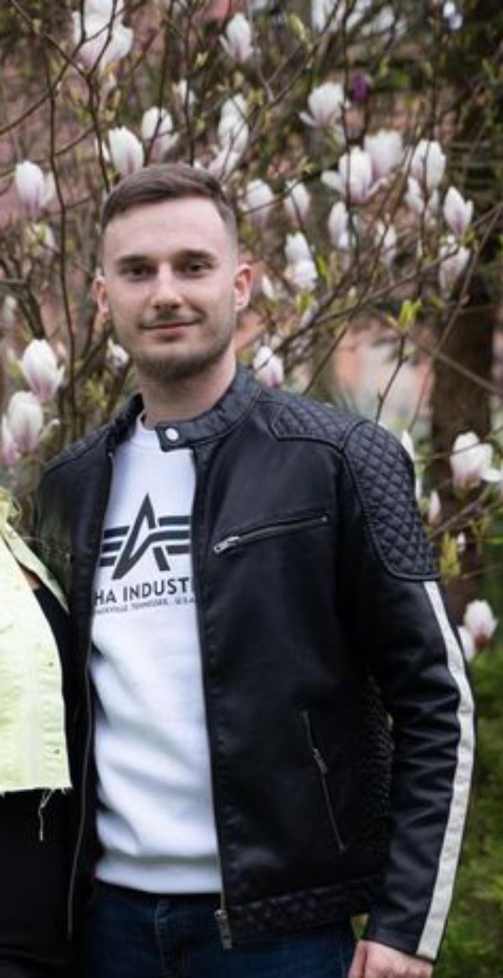 Młody mężczyzna w czarnej skórzanej kurtce z pikowanymi ramionami, stojący na tle kwitnącej magnolii.