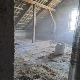 Konrad Rymarski - Poddasze w trakcie remontu: widoczne drewniane belki konstrukcyjne, ceglana ściana, okno z wpadającym światłem, worek z zaprawą oraz rozsypany materiał budowlany na podłodze.