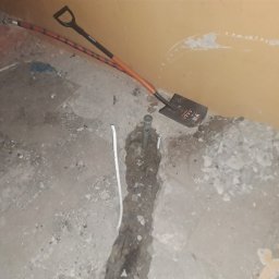 Konrad Rymarski - Wykopany rów w betonowej podłodze, widoczna rura instalacyjna, biały kabel i łopata oparta o ścianę.