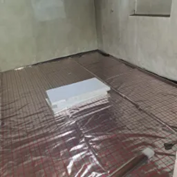 Przygotowanie do położenia podłogi: rozłożona folia grzewcza z czerwoną kratką i stos płyt izolacyjnych w rogu surowego pomieszczenia.