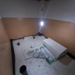 Konrad Rymarski - Przygotowanie do ocieplenia podłogi w pomieszczeniu: rozłożone płyty styropianowe z widocznymi rurami kanalizacyjnymi, zabezpieczone folią przy ścianach.