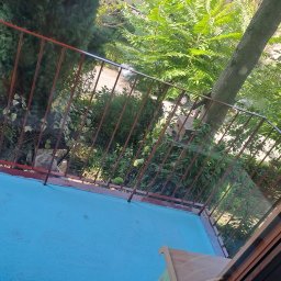 Konrad Rymarski - Widok z balkonu z turkusową podłogą i czerwoną barierką, na zewnątrz bujna zieleń drzew i krzewów.