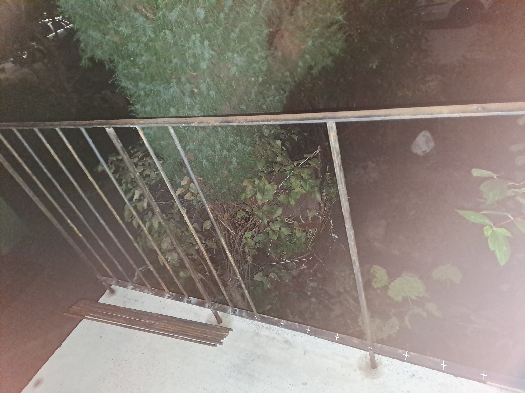 Metalowa balustrada balkonu w trakcie montażu, widoczne spawane elementy i przygotowane pręty, zarośnięte tło ogrodu.