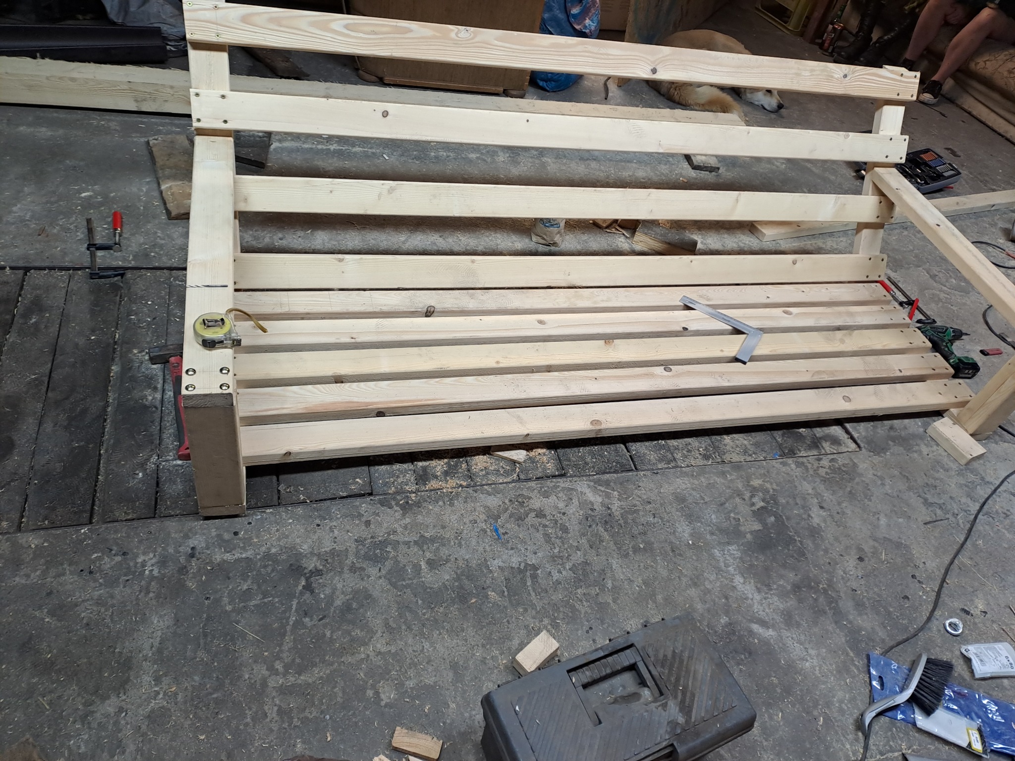 Konstrukcja z surowego drewna, prawdopodobnie rama łóżka, w trakcie montażu w warsztacie, widoczne narzędzia i akcesoria montażowe, w tle leżący pies.