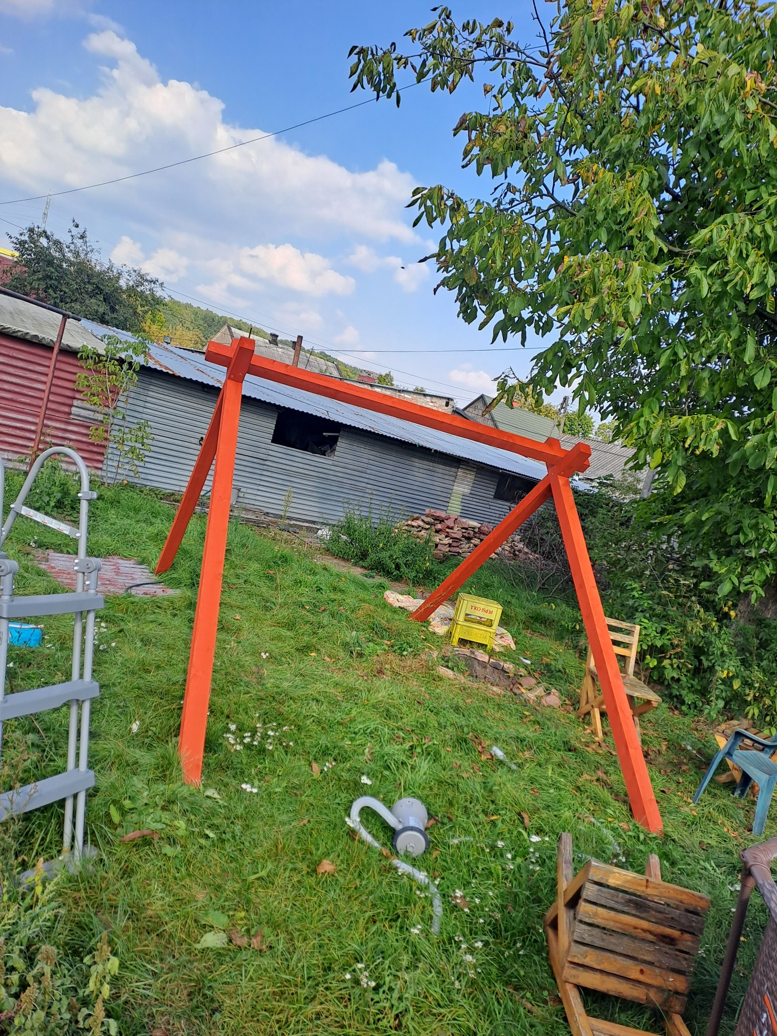 Pomarańczowa konstrukcja huśtawki w trakcie montażu na nierównym, zielonym trawniku z widocznymi elementami otoczenia, takimi jak budynek gospodarczy z blachy falistej i drzewa.