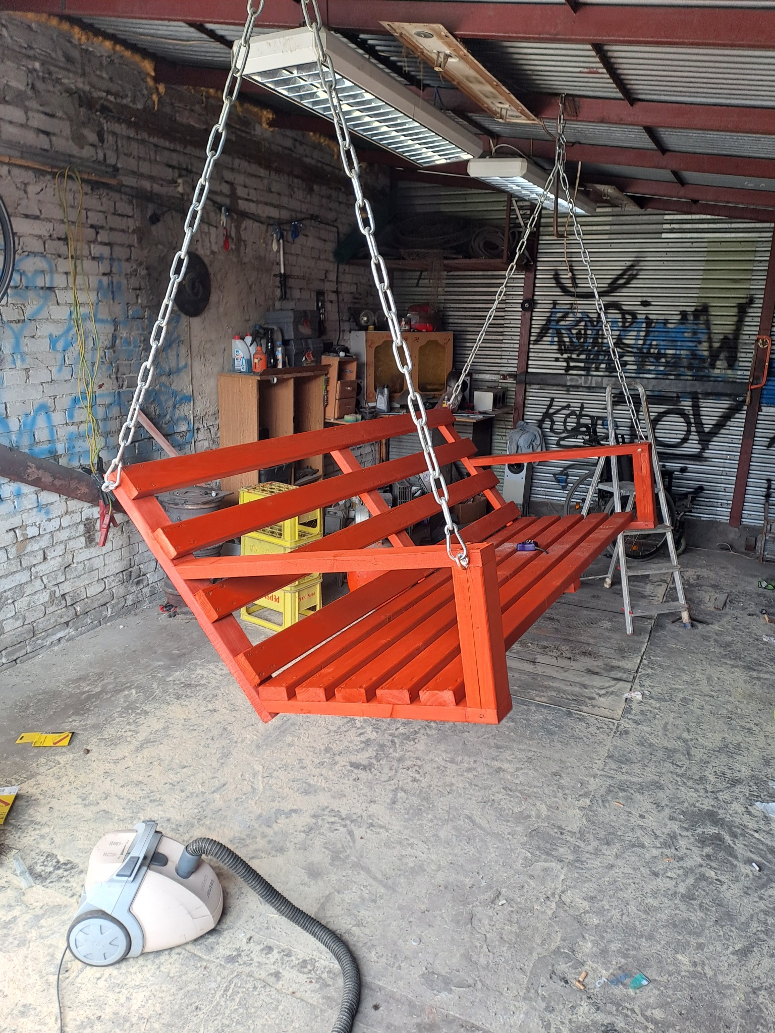 Pomarańczowa, drewniana huśtawka zawieszona na łańcuchach w warsztacie, widoczny odkurzacz, drabina i graffiti na ścianie.