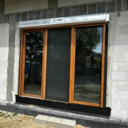 Nowe trzyskrzydłowe okno z brązowymi ramami i moskitierą, zamontowane w surowej ścianie z betonu komórkowego, widoczna roleta zwinięta w aluminiowej kasecie z logo producenta.