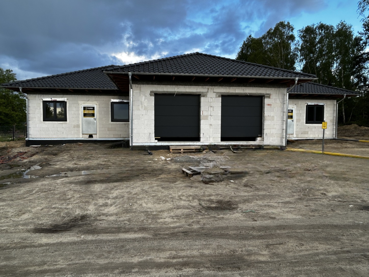 Surowy stan budowy parterowego domu z dwoma czarnymi bramami garażowymi segmentowymi, widoczne bloczki betonowe i tymczasowe zabezpieczenia budowy.