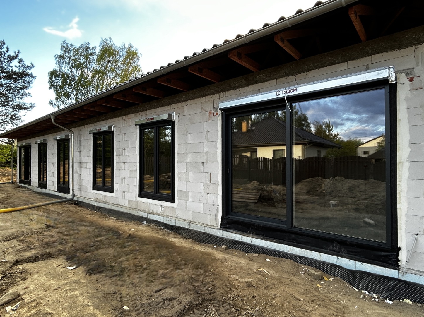 Surowy stan budynku z czarnymi oknami i drzwiami przesuwnymi, widoczne przygotowania do montażu rolet zewnętrznych, ziemia wokół budynku.