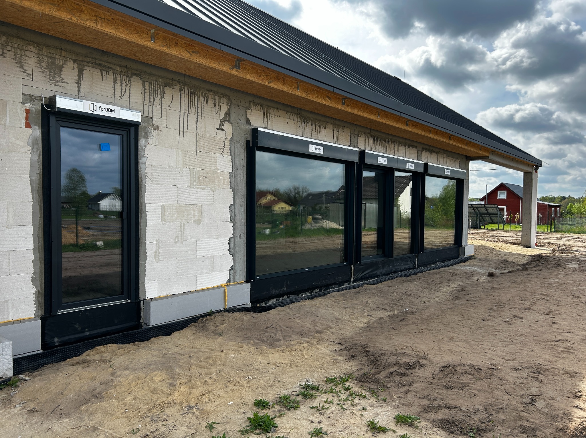 Surowy stan elewacji domu z zamontowanymi oknami i roletami zewnętrznymi w czarnych ramach, widoczne elementy izolacji fundamentów i ślady po montażu, ziemia wokół budynku.