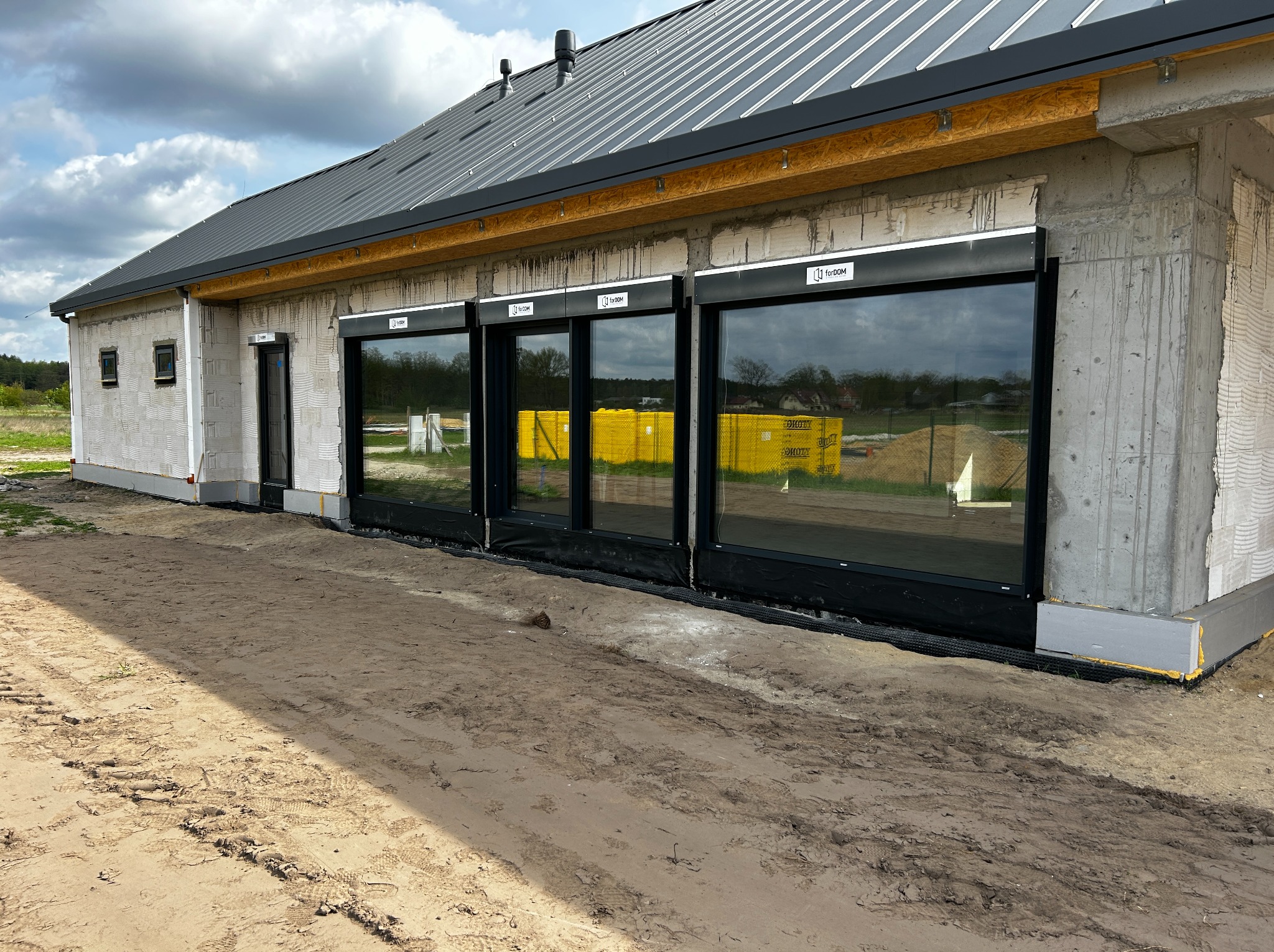 Surowy stan budynku z zamontowanymi oknami z roletami zewnętrznymi, widoczne palety z żółtą kostką brukową odbijające się w szybach, teren wokół w stanie budowy.