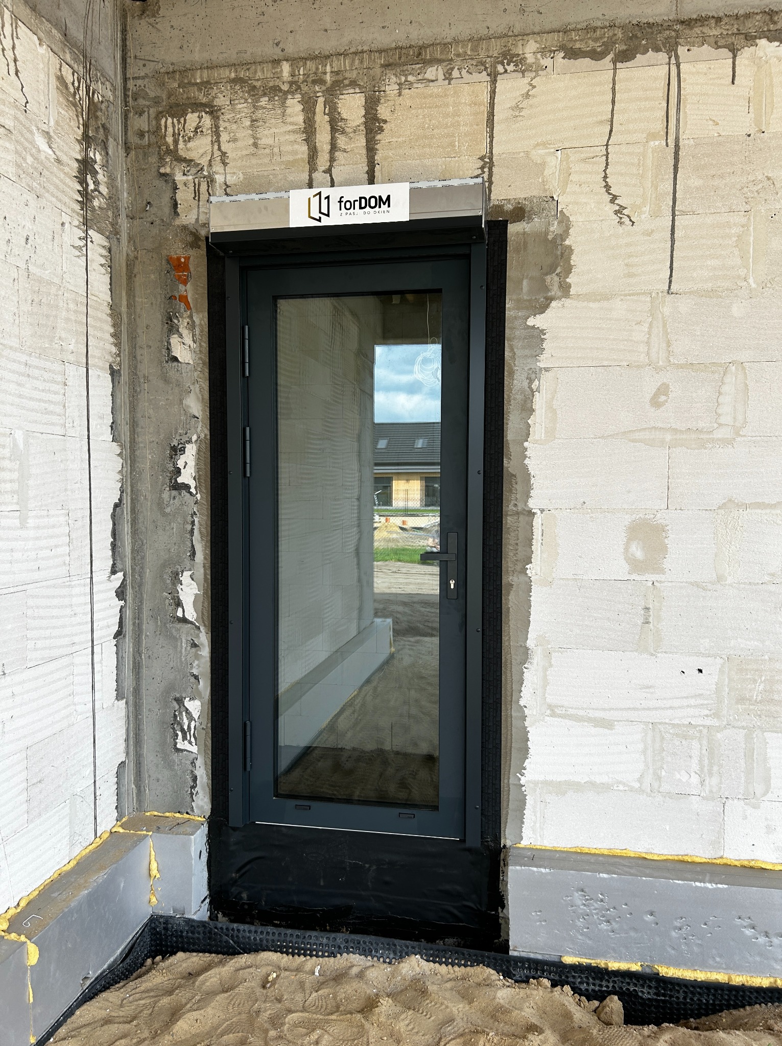 Montaż szarych drzwi zewnętrznych z szybą, widoczne surowe wykończenie budynku z betonu komórkowego, logo firmy na górze