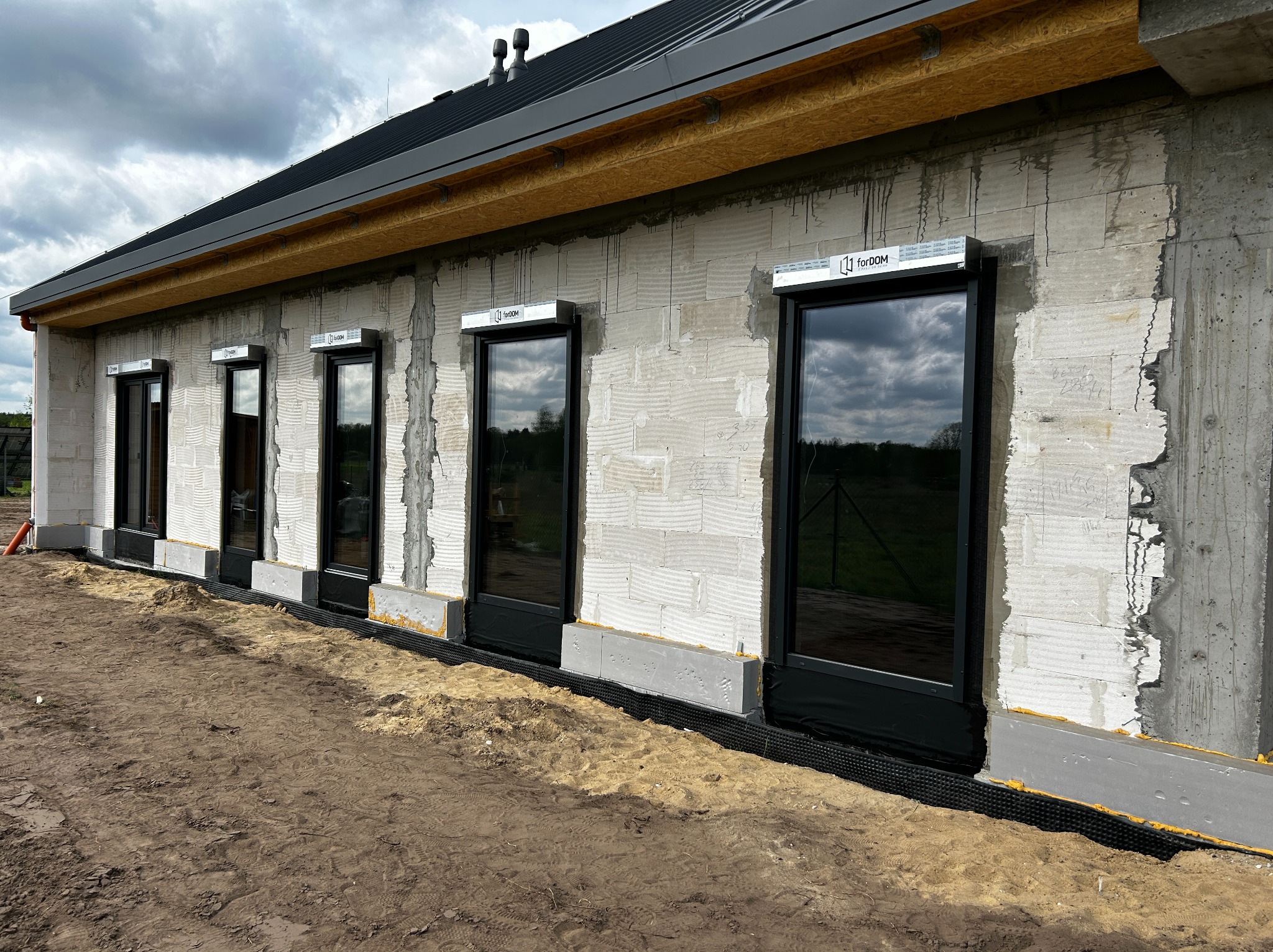 Montaż czarnych okien z roletami zewnętrznymi w surowym stanie budynku z betonu komórkowego, widoczna warstwa izolacji poziomej przy gruncie.