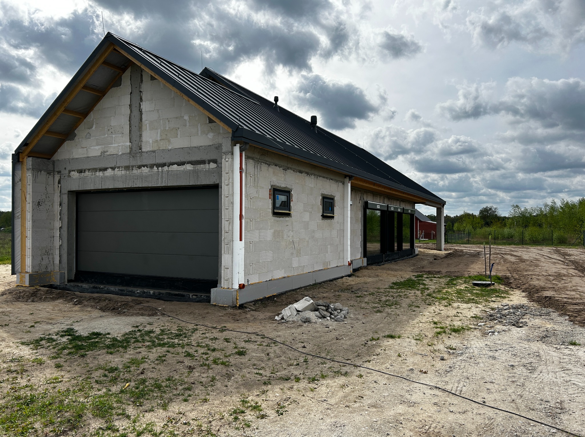 Niedokończony budynek garażu z szarą bramą garażową segmentową, widoczne bloczki betonowe, czarna blacha dachowa i przygotowane otwory okienne z czarnymi ramami, teren wokół budynku...