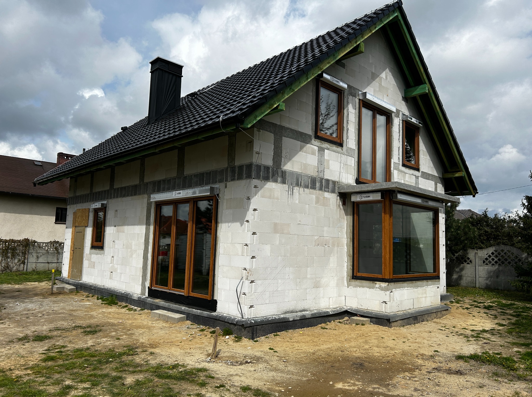 Dom w trakcie budowy z zamontowanymi oknami w drewnianej ramie, widoczna surowa elewacja z betonu komórkowego i czarna dachówka.