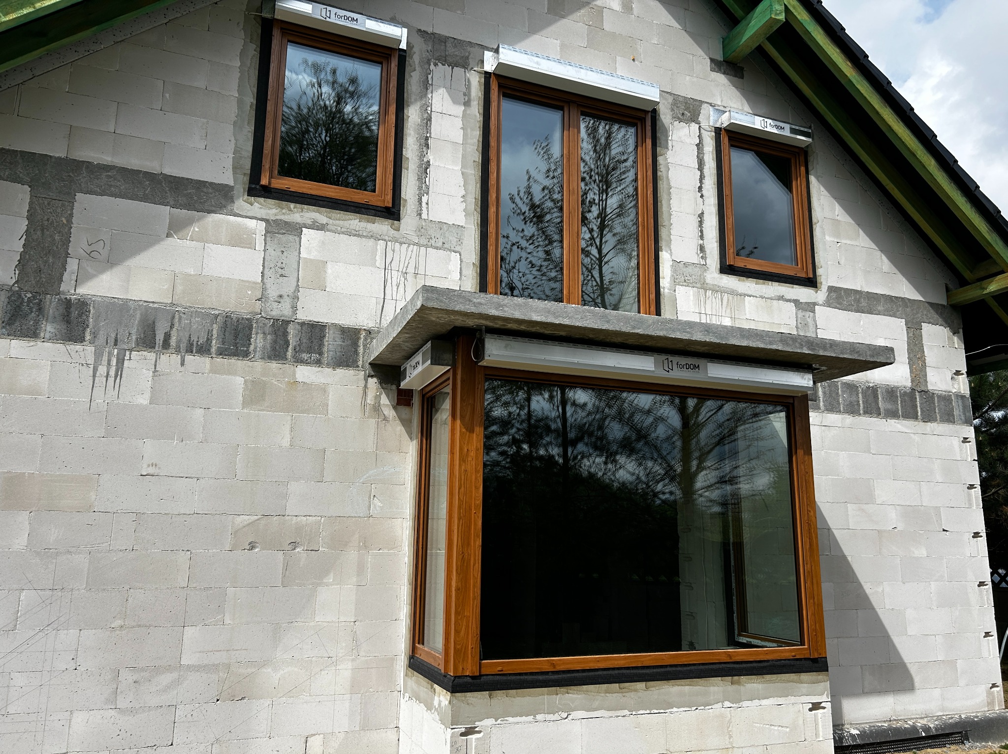 Surowa ściana budynku w trakcie budowy z zamontowanymi oknami w kolorze drewna i zwiniętymi roletami zewnętrznymi.