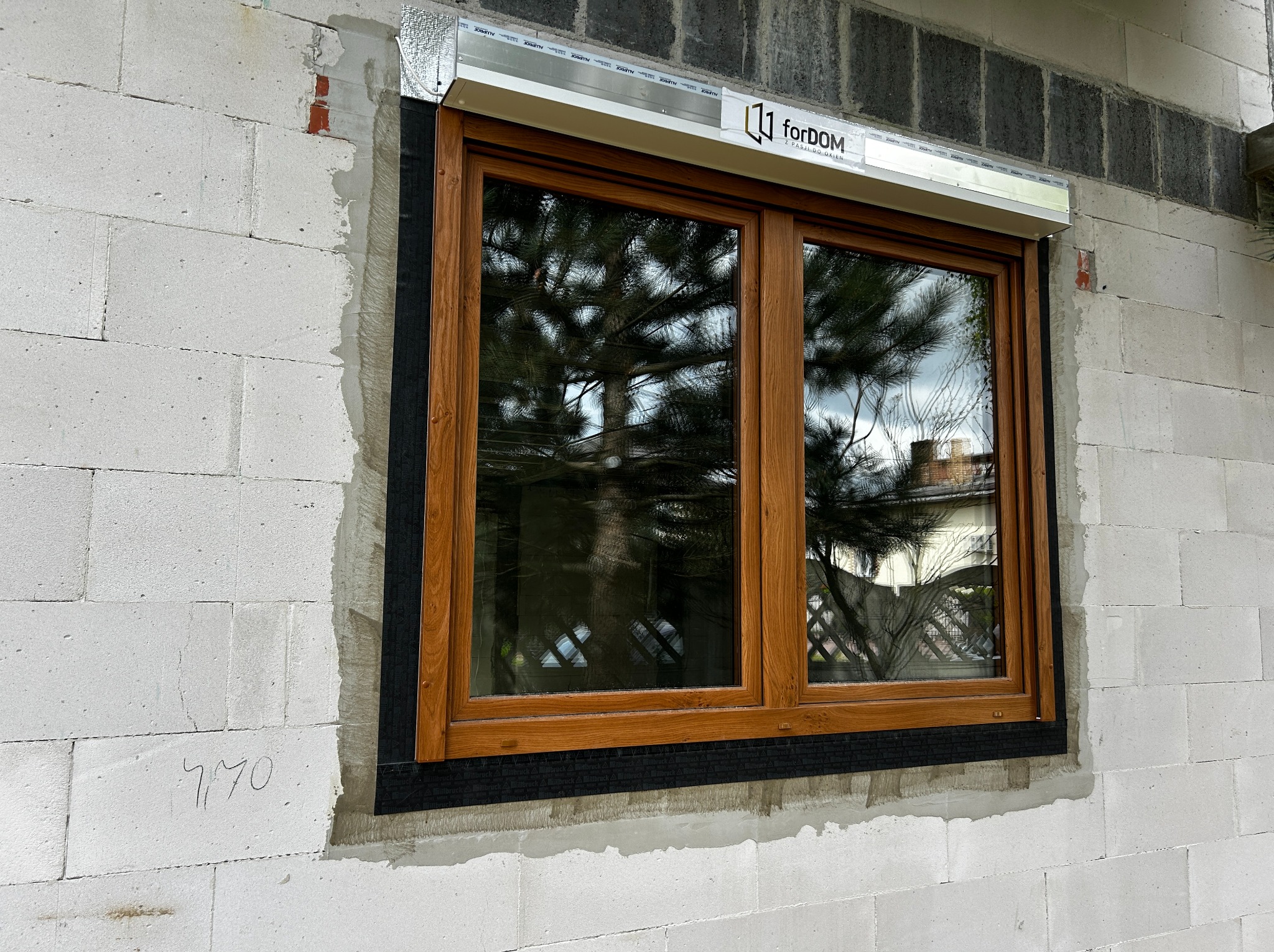 Montaż okna z roletą zewnętrzną w surowym murze z betonu komórkowego; widoczne logo producenta rolety i odbicie drzewa w szybie okna.