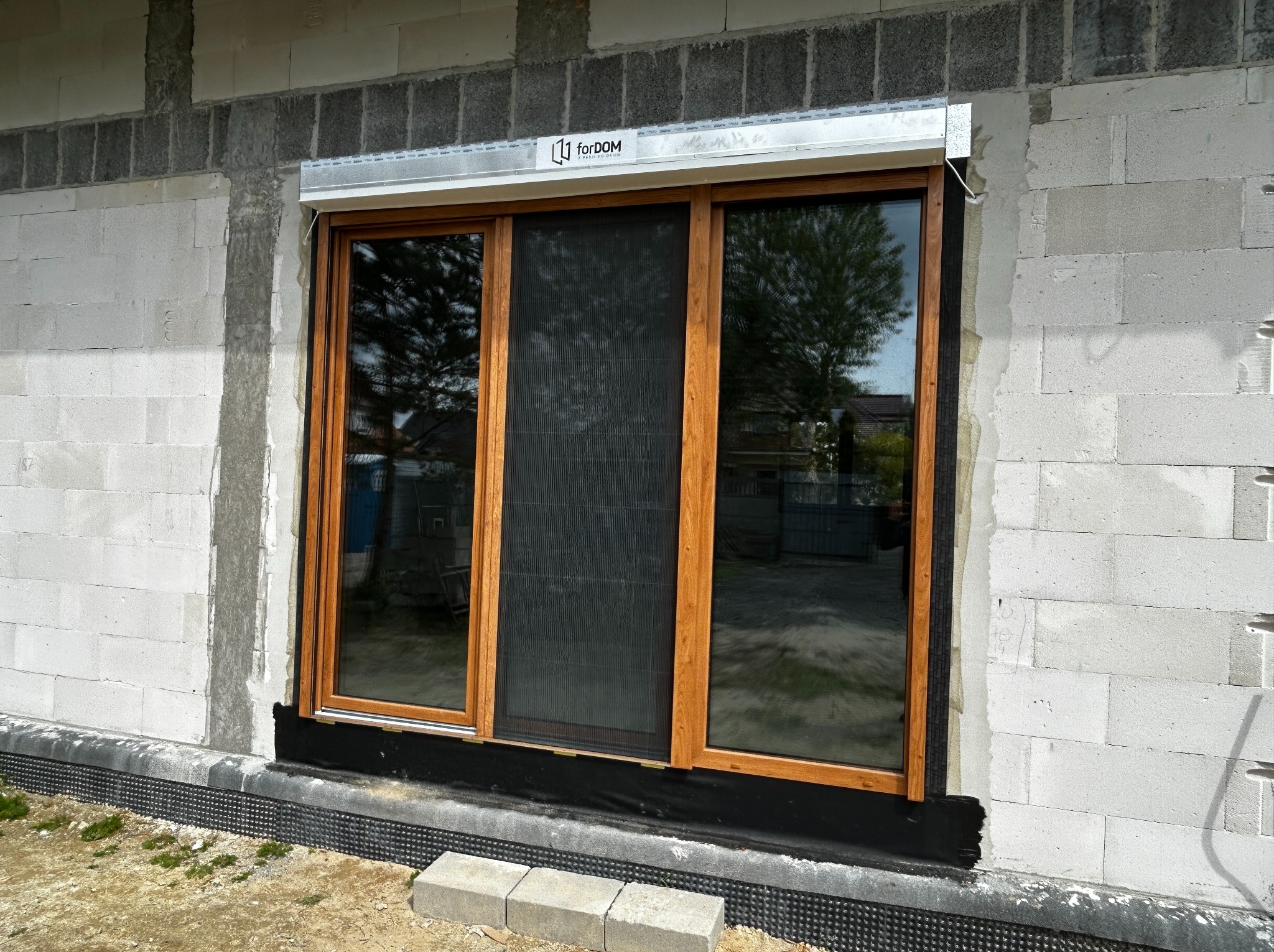 Nowe trzyskrzydłowe okno z brązowymi ramami i moskitierą, zamontowane w surowej ścianie z betonu komórkowego, widoczna roleta zwinięta w aluminiowej kasecie z logo producenta.