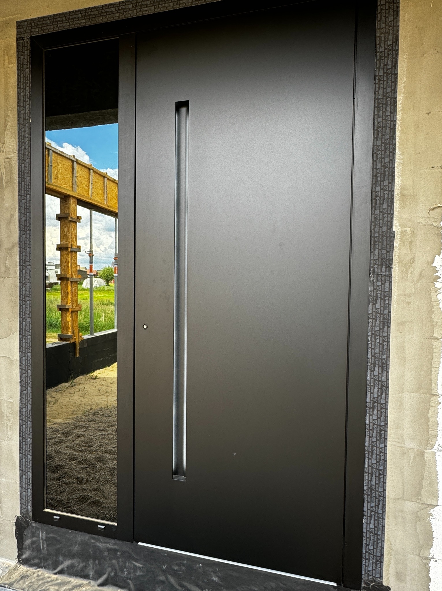 Nowoczesne, czarne drzwi wejściowe z długim, pionowym uchwytem, zamontowane w surowym stanie budynku, z widoczną konstrukcją szkieletową w odbiciu szyby.