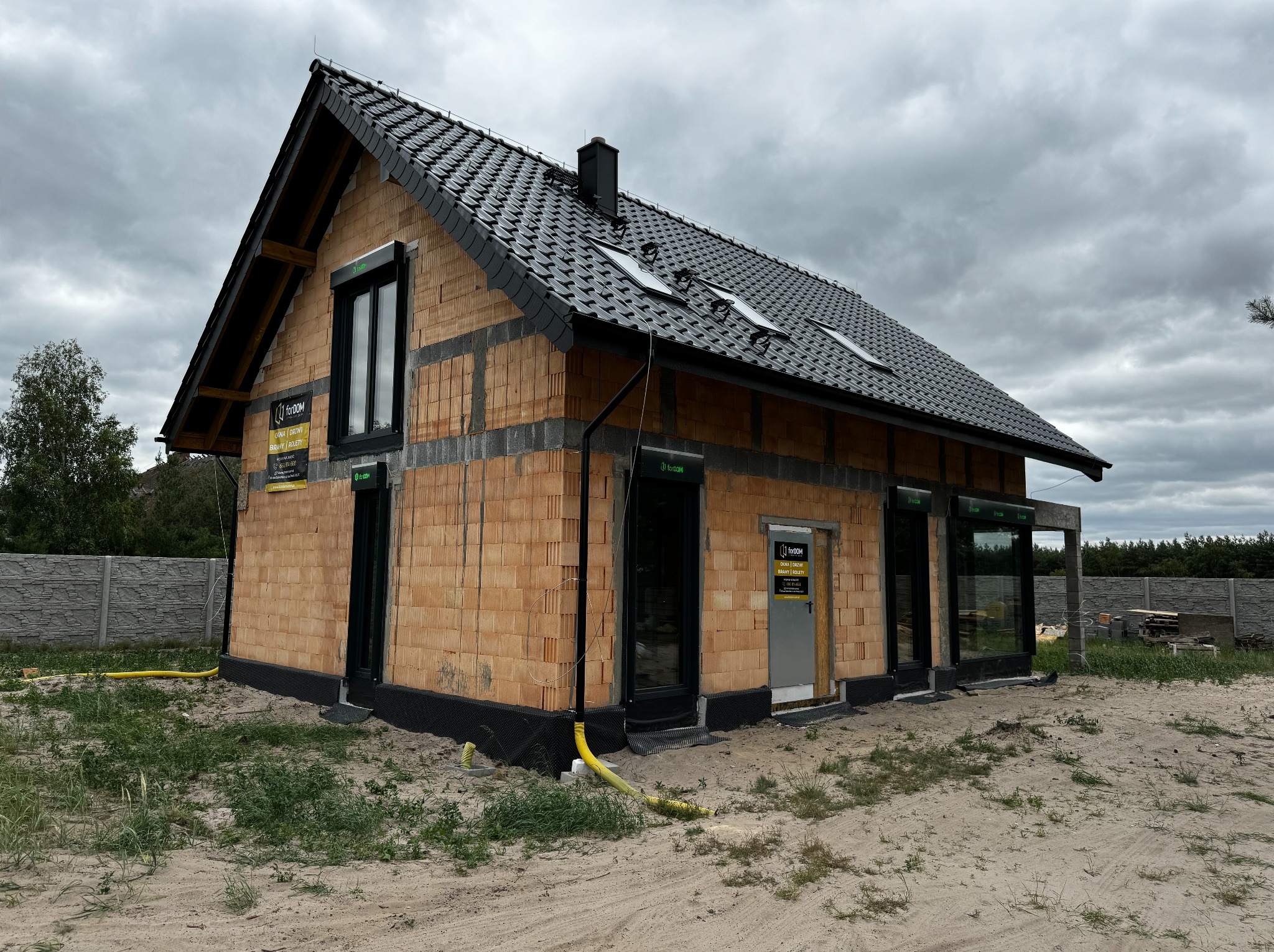 Surowy stan budynku jednorodzinnego z zamontowanymi oknami i drzwiami w czarnych ramach, widoczne także rolety zewnętrzne, ceglana elewacja w trakcie budowy, żółte rury drenażowe przy fundamentach.
