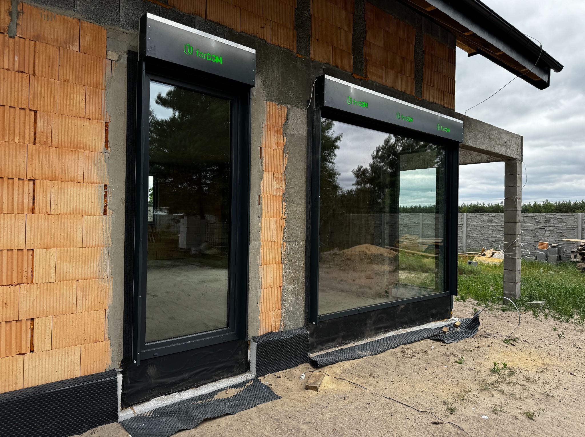 Montaż okien z roletami zewnętrznymi w surowym budynku z cegły, widoczne logo producenta na kasecie rolety, teren wokół budynku w trakcie prac ziemnych.