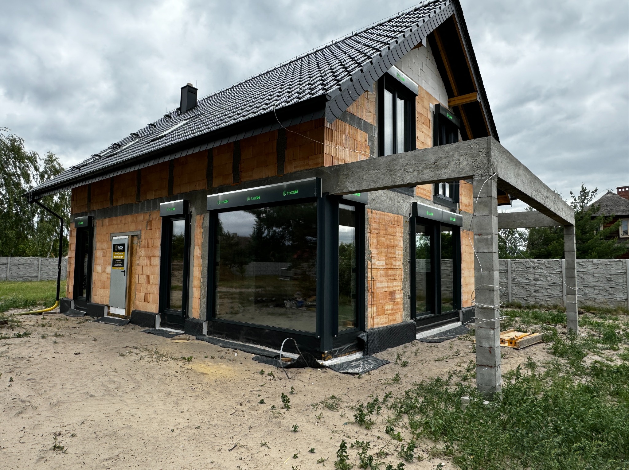 Budowa domu jednorodzinnego w trakcie montażu stolarki okiennej i drzwiowej, widoczne czarne ramy okien i drzwi, ściany z czerwonej cegły, betonowa konstrukcja zadaszenia tarasu, szara dachówka...