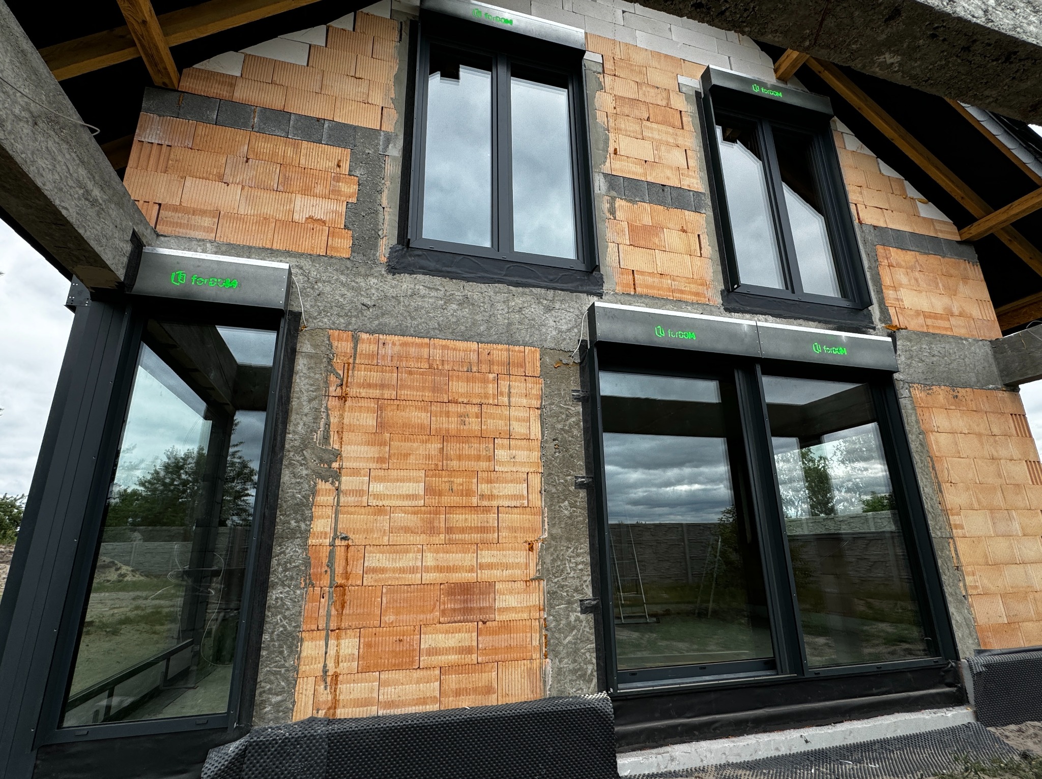 Surowy stan budynku w trakcie montażu czarnych okien z roletami zewnętrznymi, widoczne cegły ceramiczne, drewniana konstrukcja dachu i logo producenta na roletach.