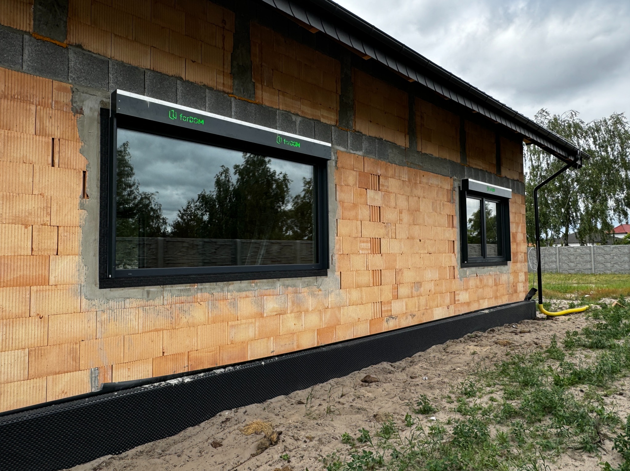 Montaż okien z roletami zewnętrznymi na surowej ścianie domu z bloczków, widoczna membrana izolacyjna przy gruncie i żółta rura odprowadzająca wodę.