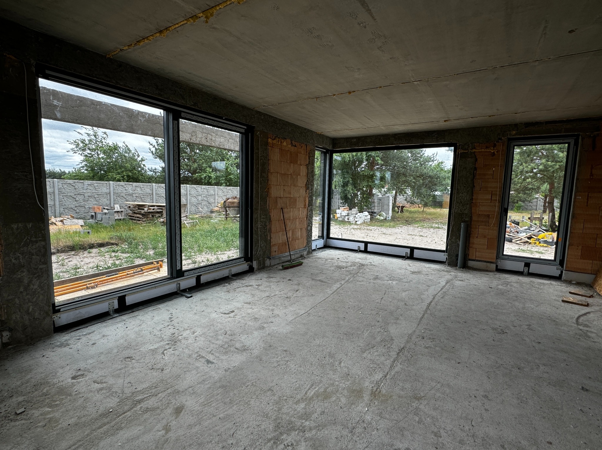 Wnętrze surowego domu z trzema dużymi, czarnymi oknami z widokiem na zewnątrz: ogród, płot i drzewa; widoczne niedokończone ściany z cegły i betonowa podłoga oraz sufit.