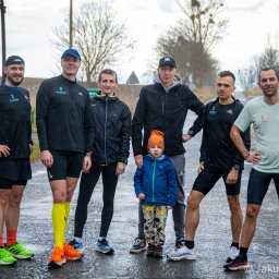 Zalewski Performance Academy - Grupa sześciu mężczyzn i chłopiec stojący na mokrej drodze, ubrani w odzież sportową, prawdopodobnie po treningu biegowym, w tle pole i drzewa.