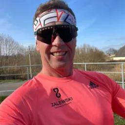 Uśmiechnięty mężczyzna w okularach przeciwsłonecznych i opasce na głowie, w różowej koszulce z logo 'ZALEWSKI Performance Academy' i 'Adidas', pozujący na tle krajobrazu z drzewami i błękitnym niebem.