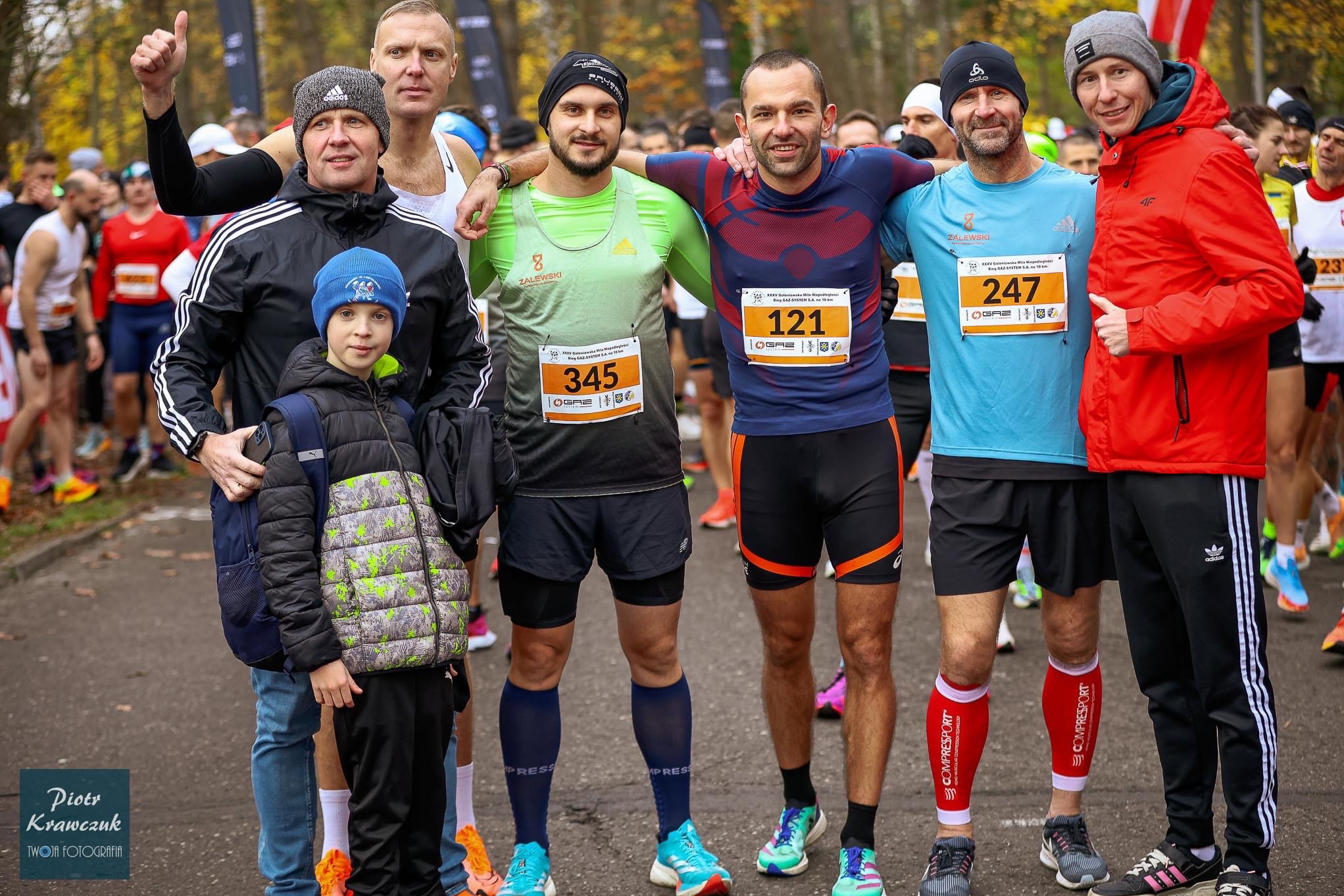 Grupa biegaczy pozuje do zdjęcia przed biegiem, widoczne numery startowe, odzież sportowa i uśmiechy na twarzach, w tle tłum uczestników i drzewa.
