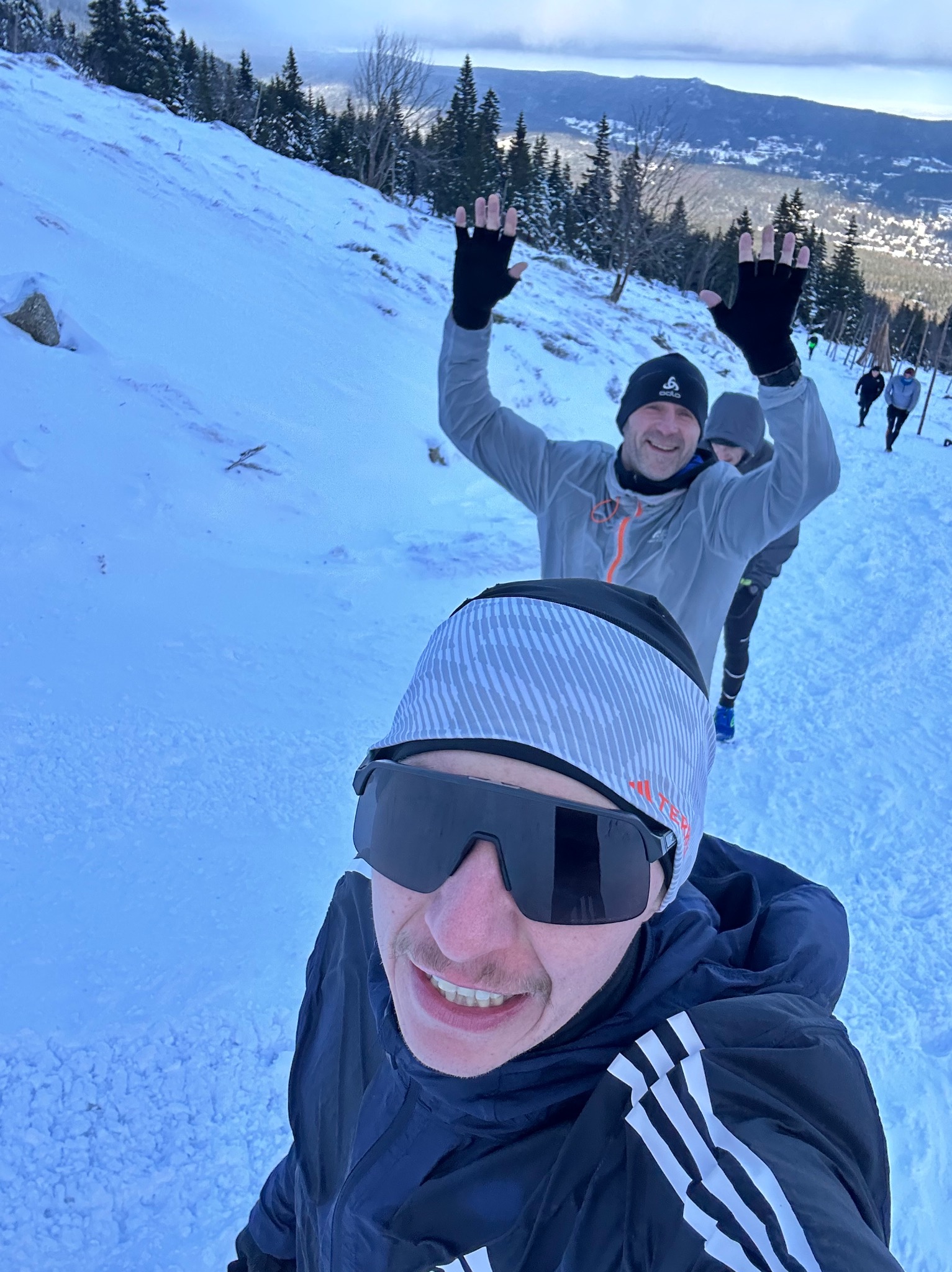 Grupa osób biegnie pod górę po śniegu; selfie mężczyzny w okularach przeciwsłonecznych na pierwszym planie, w tle widoczny mężczyzna z uniesionymi rękami i panorama miasta w oddali.