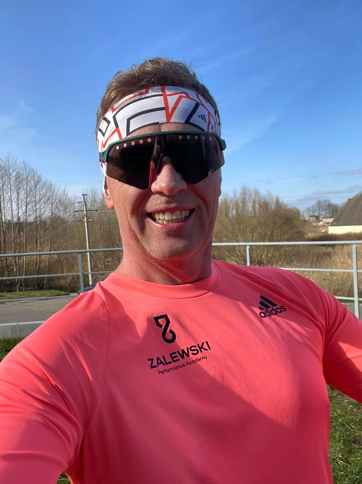 Uśmiechnięty mężczyzna w okularach przeciwsłonecznych i opasce na głowie, w różowej koszulce z logo 'ZALEWSKI Performance Academy' i 'Adidas', pozujący na tle krajobrazu z drzewami i błękitnym niebem.