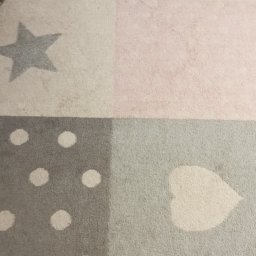 Monika Kwiran - Fragment dywanu dziecięcego w pastelowych kolorach z wzorami gwiazdy, kropek i serca, widoczne ślady użytkowania.