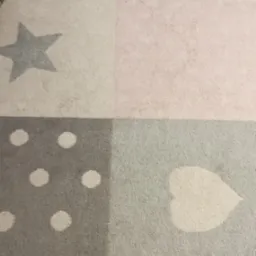 Fragment dywanu dziecięcego w pastelowych kolorach z wzorami gwiazdy, kropek i serca, widoczne ślady użytkowania.