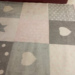 Monika Kwiran - Fragment wzorzystego dywanu w odcieniach szarości, różu i bieli z motywami serc, gwiazd i kropek, leżącego na podłodze.