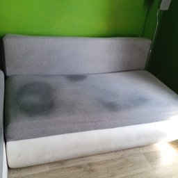 Monika Kwiran - Szara kanapa z wyraźnymi plamami na siedzisku, biały dół, na tle zielonej ściany i drewnianej podłogi.