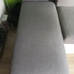 Fragment szarej kanapy modułowej z widoczną fakturą tkaniny i białym obramowaniem, obok ściana w odcieniach zieleni oraz kartonowe pudełko na podłodze.