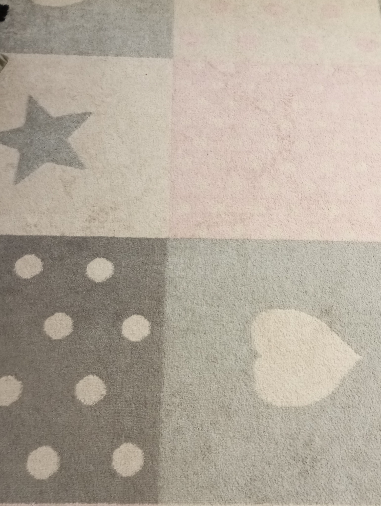 Fragment dywanu dziecięcego w pastelowych kolorach z wzorami gwiazdy, kropek i serca, widoczne ślady użytkowania.