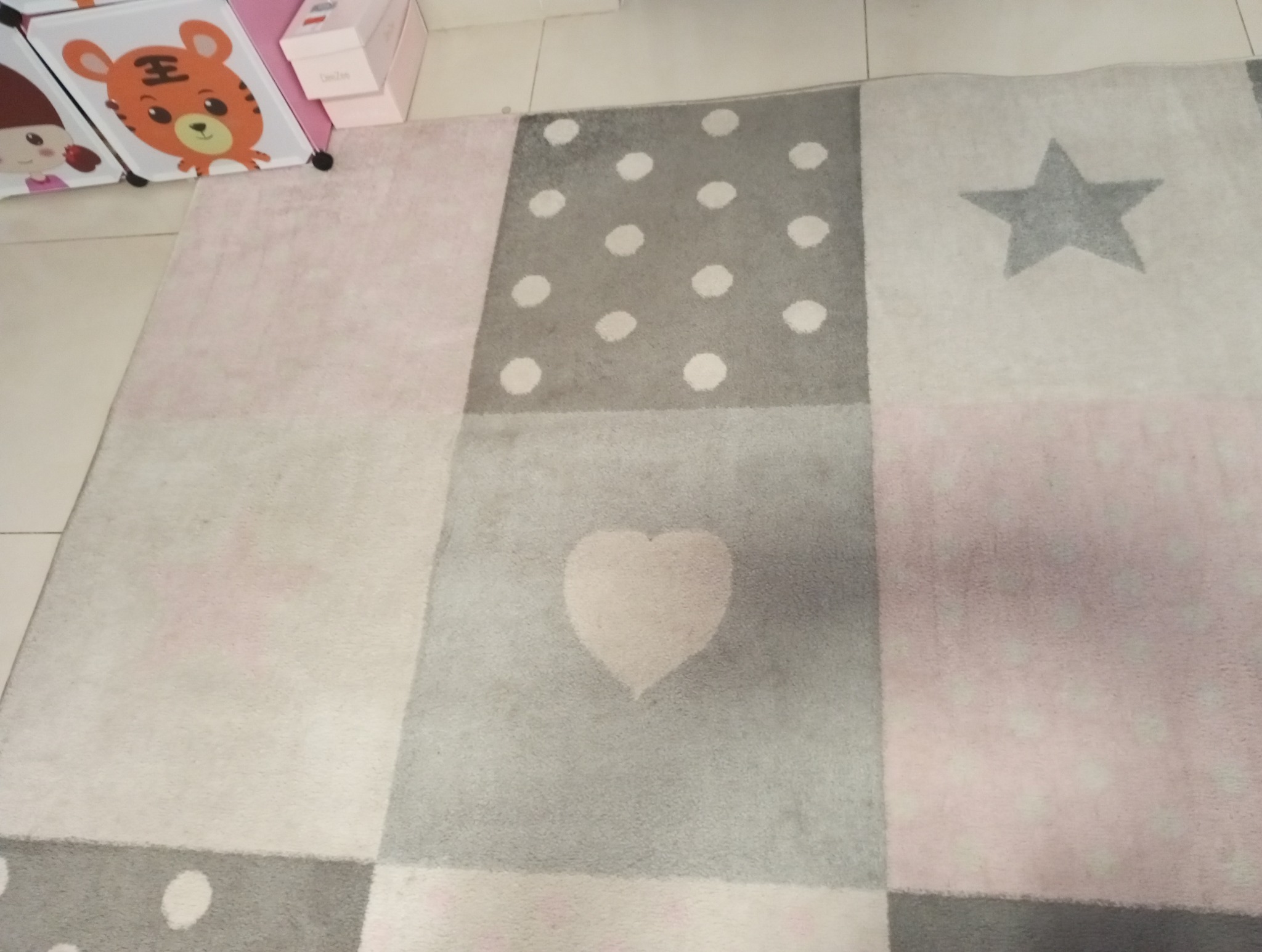 Wielobarwny dywan dziecięcy z motywem gwiazdy, serca i kropek, leżący na podłodze z jasnymi płytkami, obok szafki z motywami zwierzęcymi.