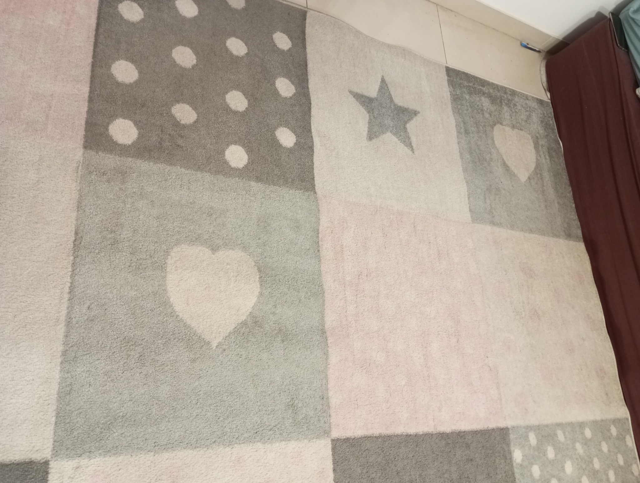 Wielobarwny dywan dziecięcy w geometryczne wzory: gwiazda, serce, kropki, w odcieniach szarości i różu, częściowo przykryty brązową zasłoną.